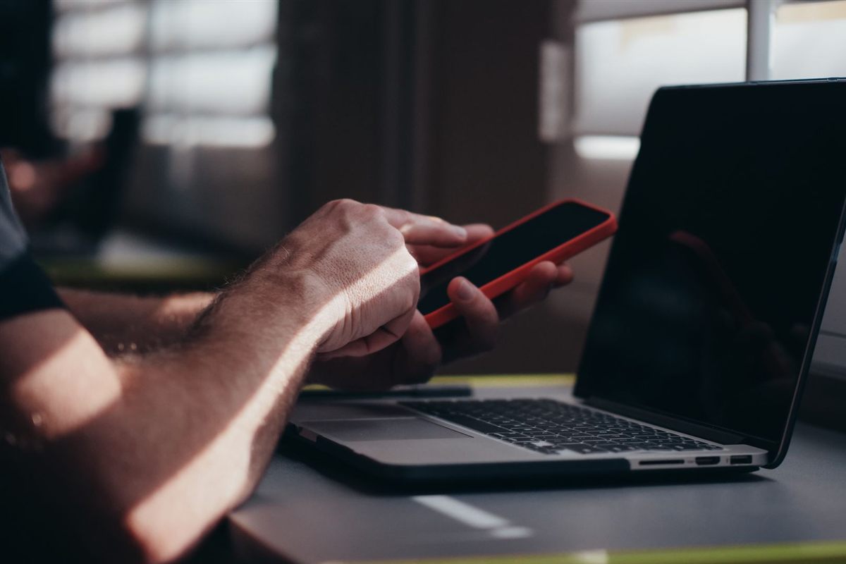 A hand holding a personal phone beside an open work laptop